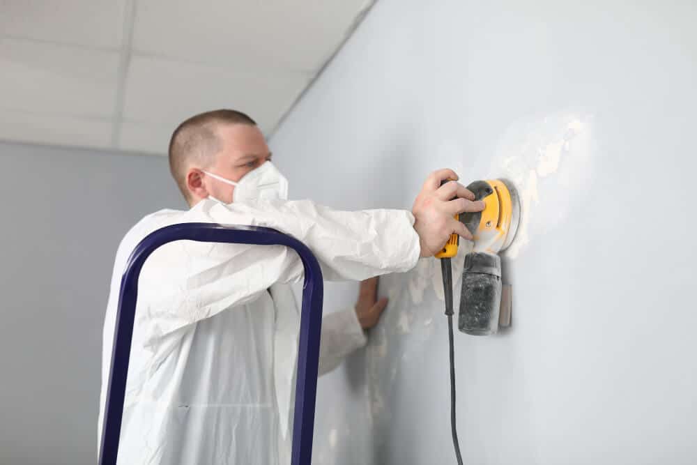 Main d'un peintre ponçant légèrement un mur blanc recouvert d'une sous-couche pour enlever les imperfections.