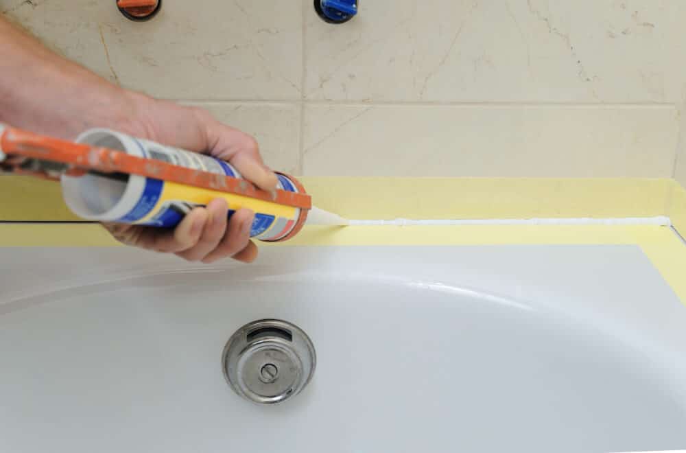 Main appliquant un joint de mastic silicone sanitaire blanc dans l'espace entre le bord d'une baignoire et un mur carrelé.